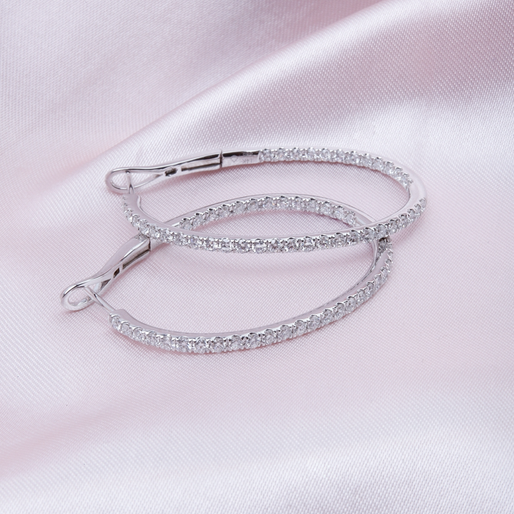 A pair of hoop earrings with a single row of round diamonds laying against a light-pink background.