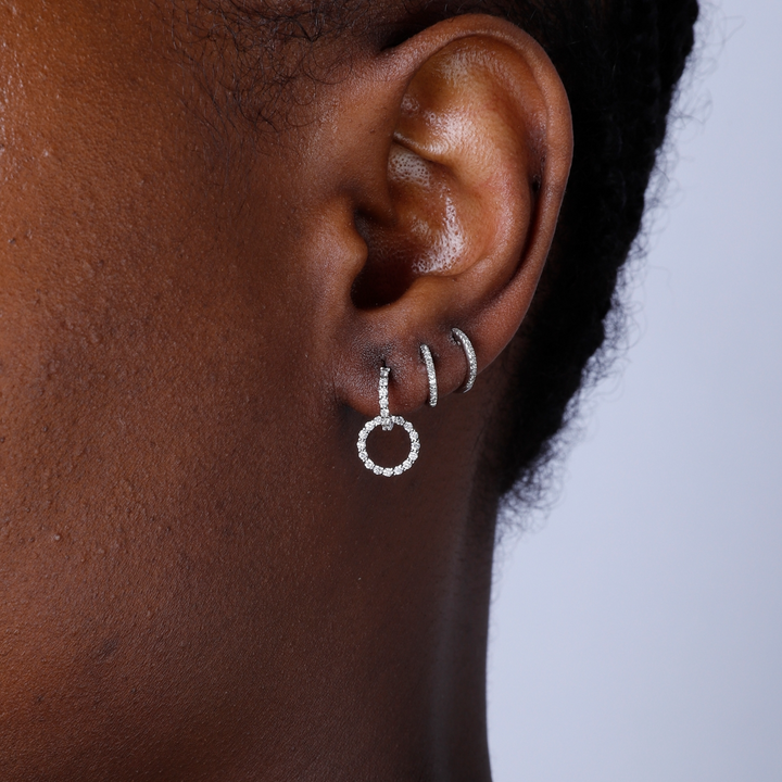 A woman with short black hair is wearing a pair of drop earrings with a diamond-set open circle design.