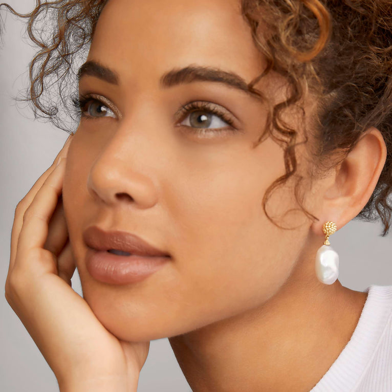 A woman wearing a white shirt and drop earrings with an 18K gold Caviar beading frame and cultured freshwater pearl baroque. 