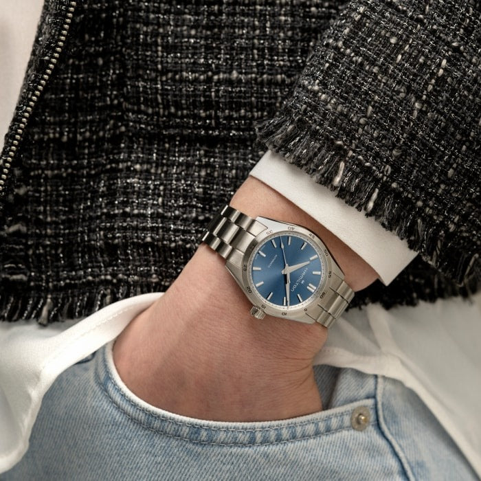 A woman wearing a black jacket and a Hamilton watch around her wrist. It features a blue dial, silver hands and markers, a stainless steel bezel, and a stainless steel bracelet.