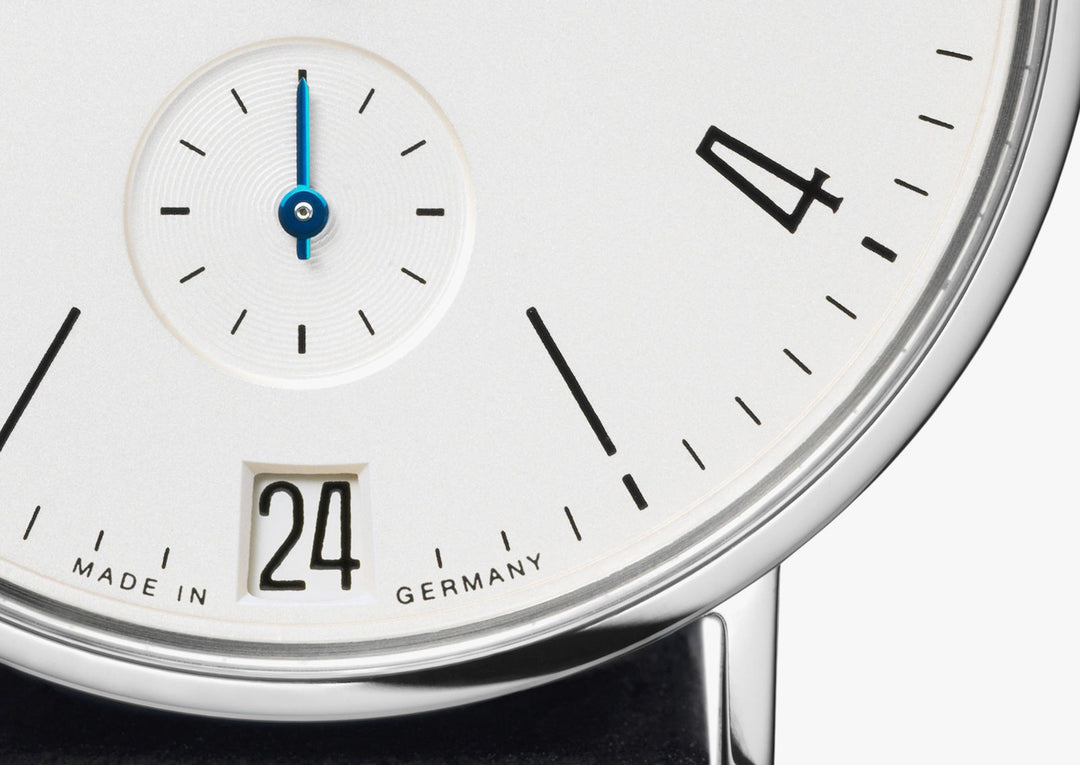 This is a close-up image of a Nomos Glashutte watch, focusing on the bottom part of the dial. It features a white dial, black & blue hands and markers, a stainless steel bezel, and a black leather strap.