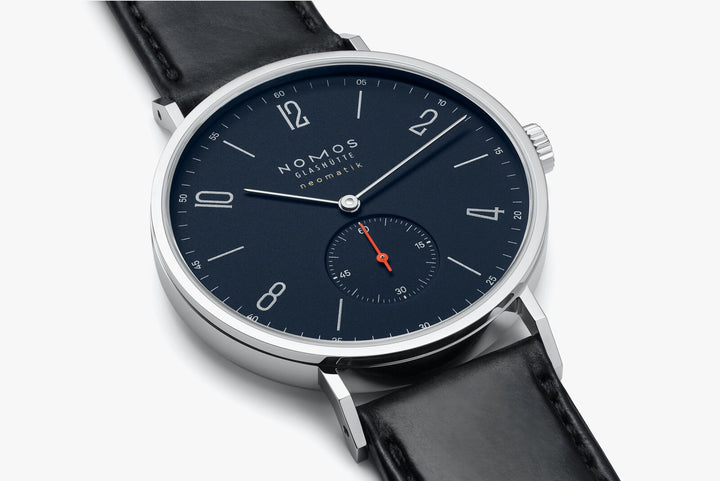 This is a close-up image of a Nomos Glashutte watch angled to the right. It features a dark blue dial, white hands and markers, a stainless steel bezel, and a black strap.