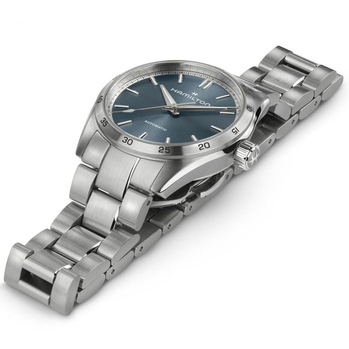 A Hamilton watch is laid out tilted to the right against a white background. It features a blue dial, silver hands and markers, a stainless steel bezel, and a stainless steel bracelet.