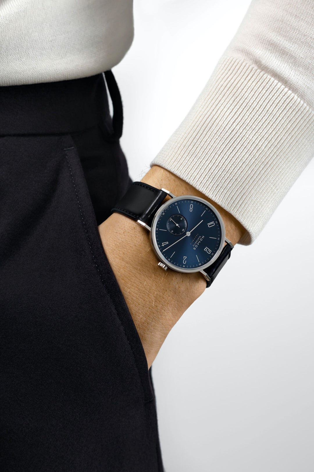 A man wearing a Nomos Glashutte watch on his wrist. It features a blue dial, silver hands and markers, a stainless steel bezel, and a black leather strap.