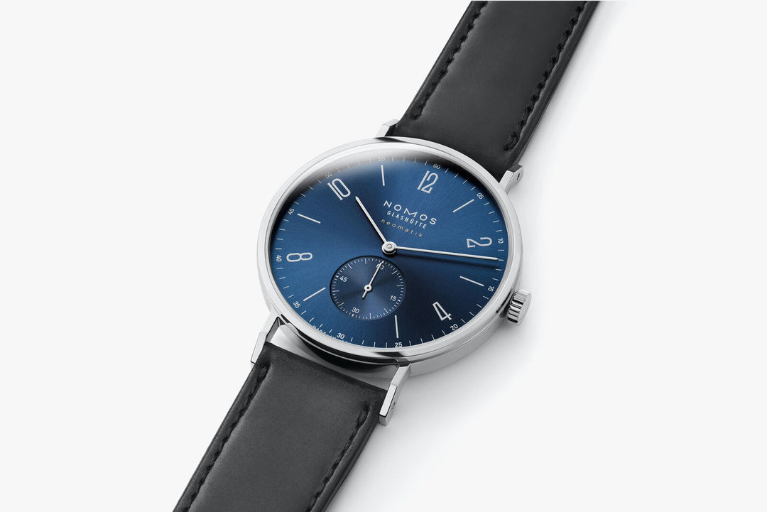 This is a close-up image of a Nomos Glashutte watch angled to the right. It features a blue dial, silver hands and markers, a stainless steel bezel, and a black leather strap.