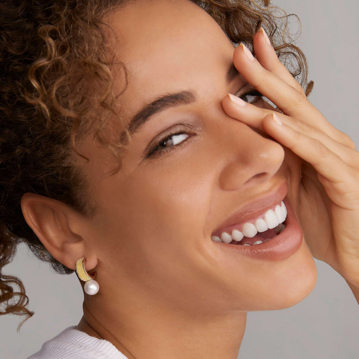 A woman is wearing a white top and a pair of hoop earrings with 18K gold and sterling silver Caviar beading with pearl drop detailing. 