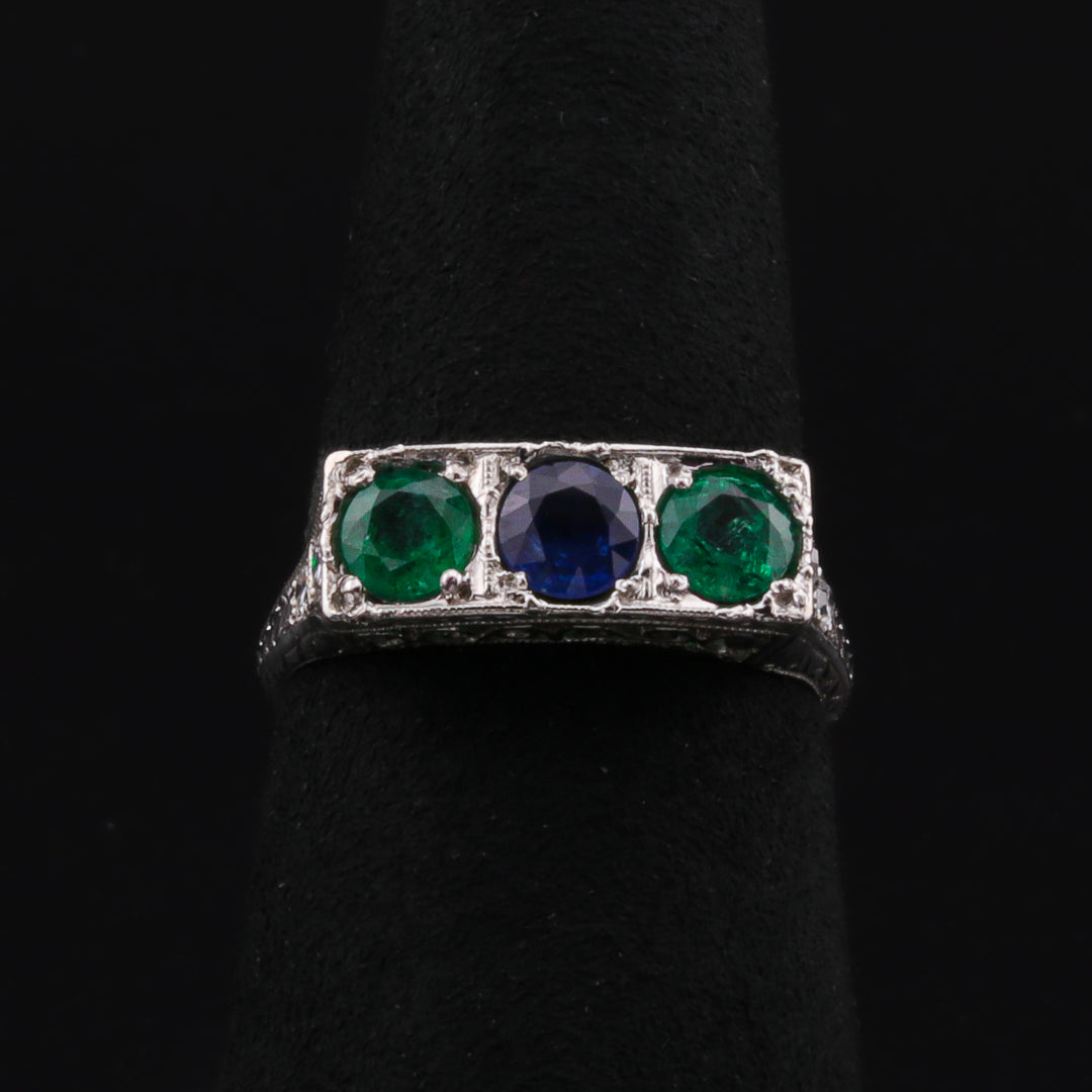 This is a close-up of a platinum ring with a trio of stunning gemstones—two vibrant emeralds and a sapphire—set in an intricate antique platinum setting. The ring is resting on a black ring holder against a black background.