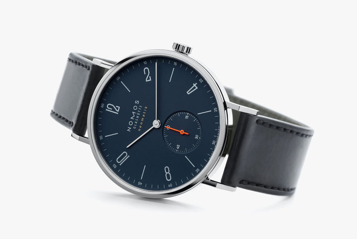 A Nomos Glashutte watch resting on the left side against a white background in the middle. It features a dark blue dial, white hands and markers, a stainless steel bezel, and a black strap.