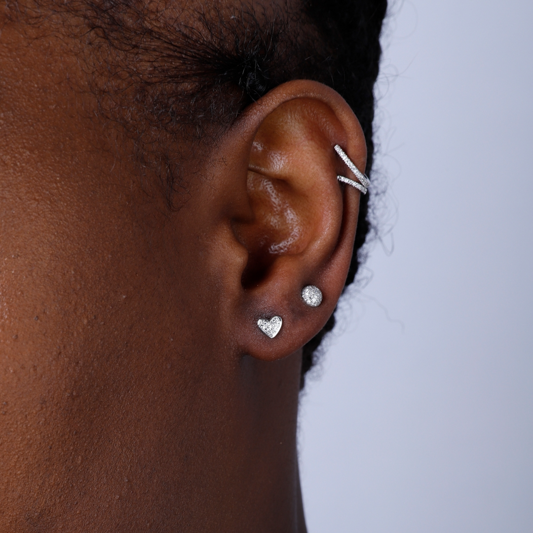 A woman with short black hair is wearing a pair of stud earrings with a heart shaped design with clustered round diamonds. 