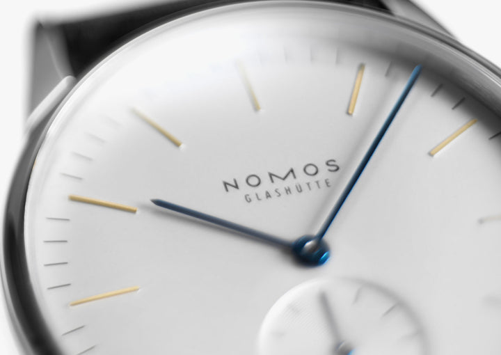 A close-up image of a Nomos Glashutte watch angled to the right, focusing on the dial. It features a white dial, gold & blue hands, and markers, and a stainless steel bezel.