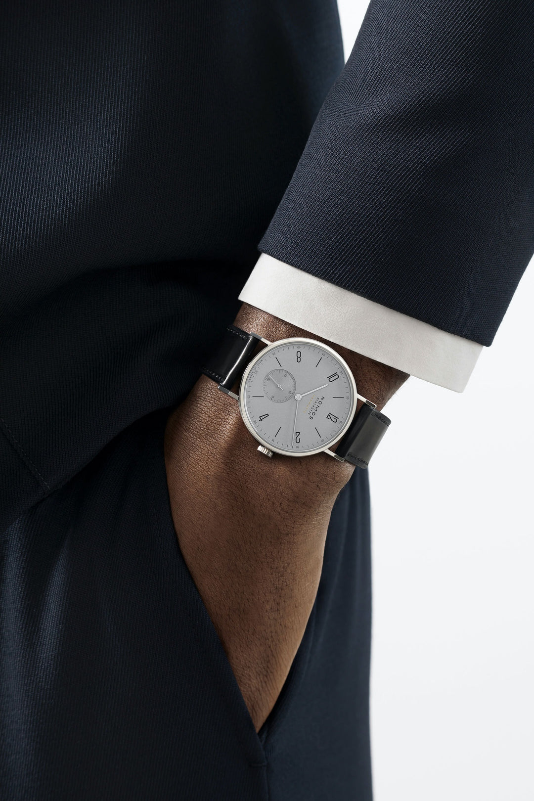 A man wearing a Nomos Glashutte watch on his wrist. It features a white dial, black and white hands and markers, a stainless steel bezel, and a black leather strap.