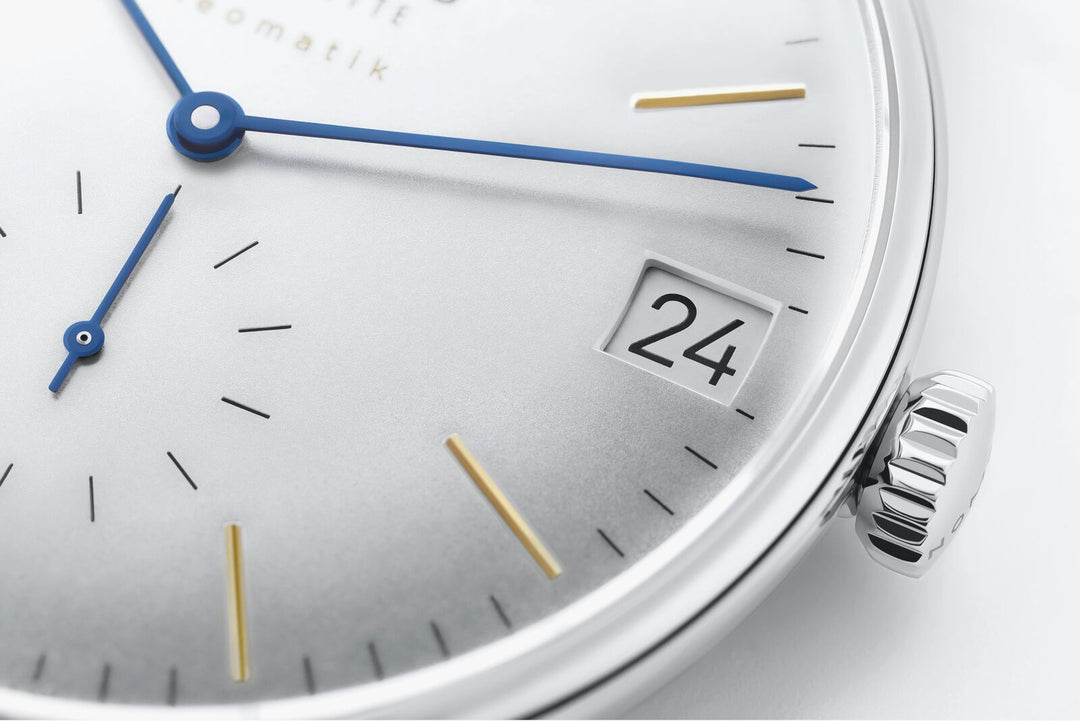 This is a close-up image of a Nomos Glashutte watch angled to the right. It features a white dial, gold & blue hands and markers, a stainless steel bezel, and a black strap.