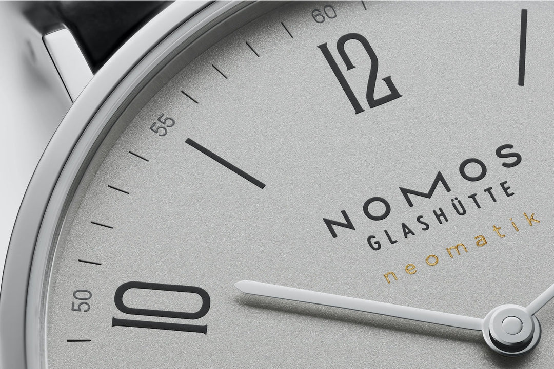 A close-up of a Nomos Glashutte watch angled to the left, focusing on the dial. It features a white dial, black and white hands and markers, a stainless steel bezel, and a black leather strap.