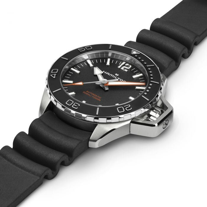 A Hamilton watch is laid out tilted to the right against a white background. It features a black dial, white hands and markers, a steel case, a black bezel, a steel crown, and a black rubber strap.