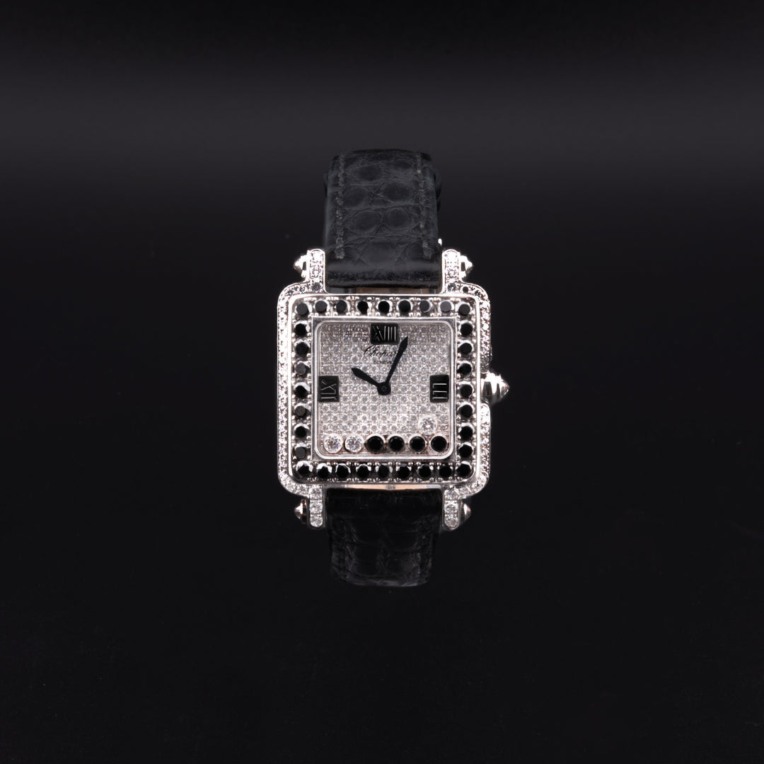 This is a close-up of a Chopard watch. The watch features a diamond set dial, a diamond case and crown, a black diamond-set bezel, black hands and markers, and a black leather strap on a black background.