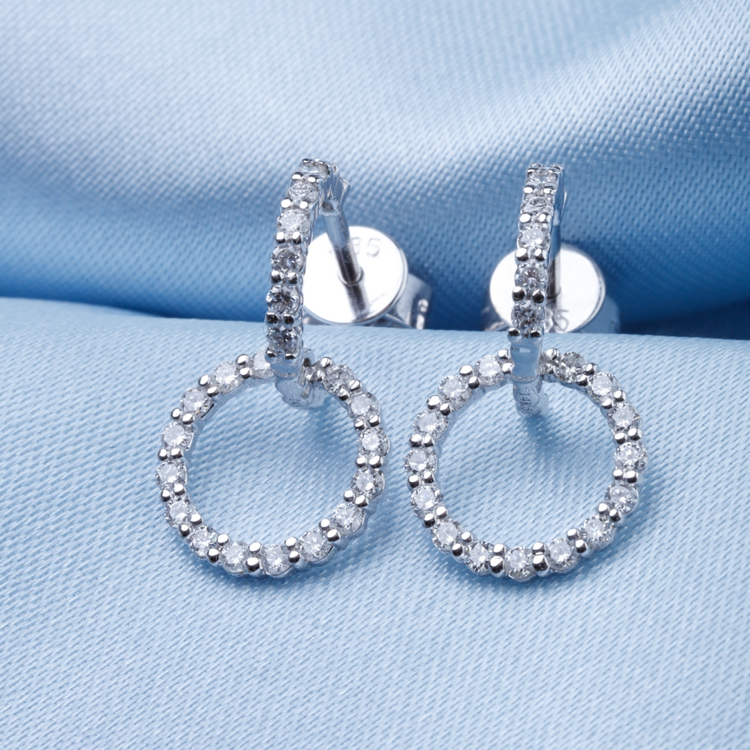 A pair of drop earrings with a diamond-set open circle design resting against a light-blue background.