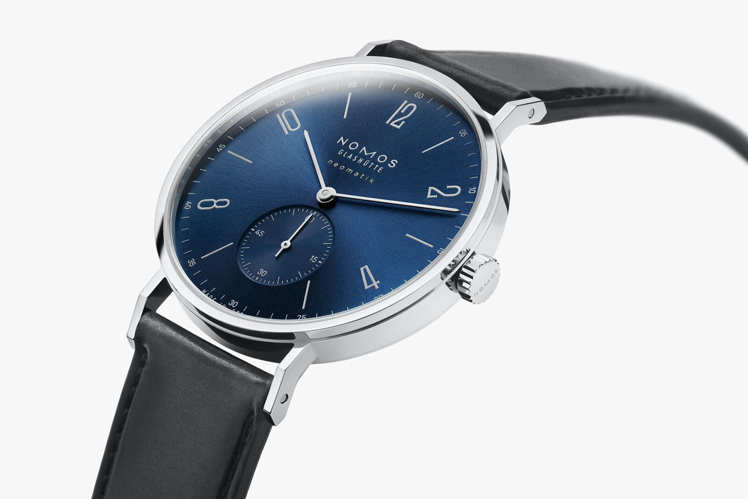 A side view of a Nomos Glashutte watch angled & curved to the left. It features a blue dial, silver hands and markers, a stainless steel bezel, and a black leather strap.