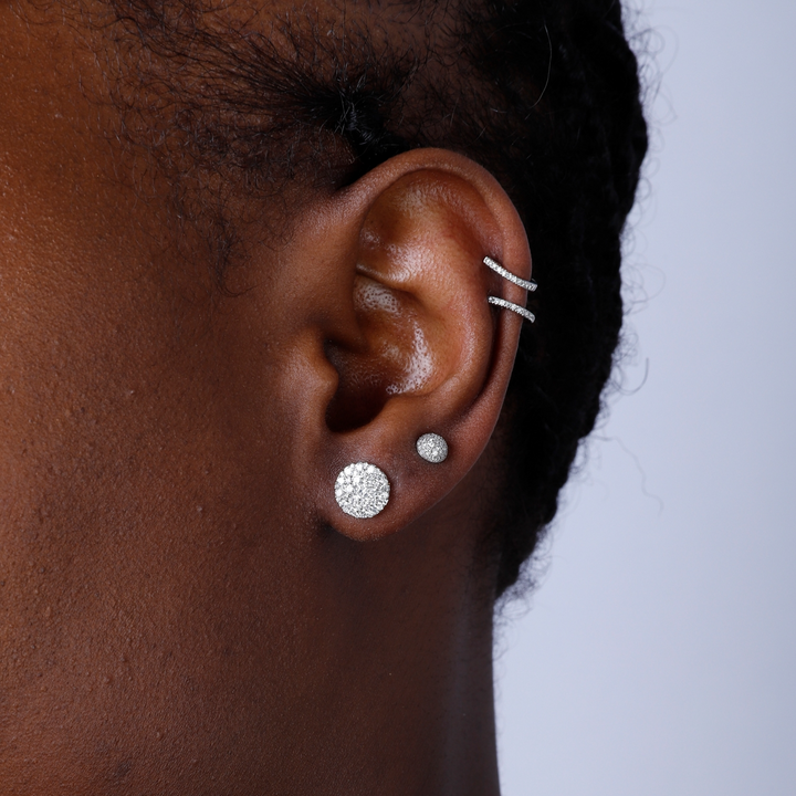 A woman with short black hair is wearing a pair of stud earrings with a round diamond shape with a cluster of diamonds.