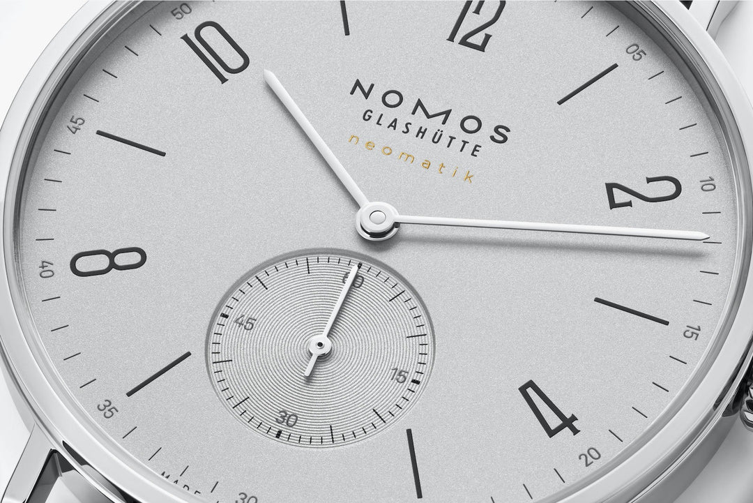 This is a close-up image of a Nomos Glashutte watch angled to the right, focusing on the dial. It features a white dial, black and white hands and markers, a stainless steel bezel, and a black leather strap.