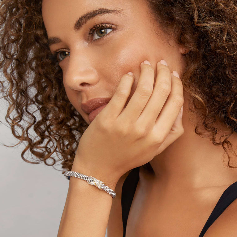 A close-up of a woman wearing a black top and a Sterling Silver & 18k gold bracelet. The bracelet features a diamond-set X station, caviar beading, and a stainless steel clasp.