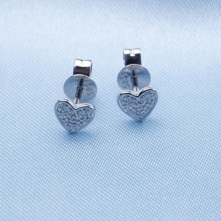 A pair of stud earrings with a heart shaped design with clustered round diamonds rests against a light-blue background.