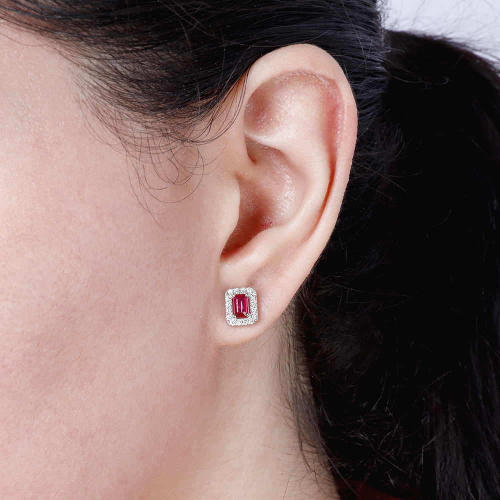 A woman with brunette hair is wearing a pair of stud earrings, a ruby emerald-shaped diamond in the middle, and a surrounding diamond frame. 