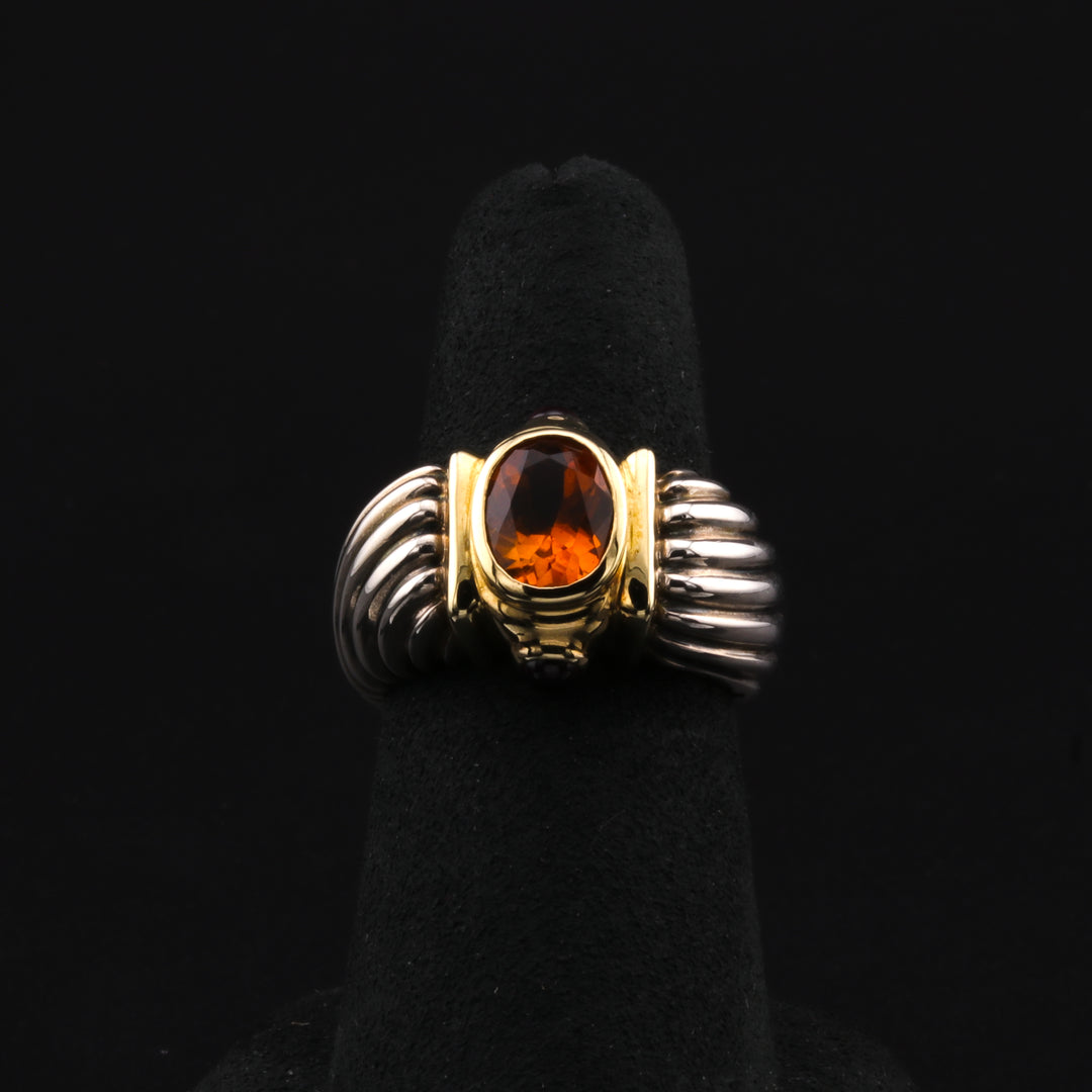 This is a close-up of a two-tone ring with a radiant citrine center stone accented by vibrant rhodolite side stones and a signature David Yurman sterling silver band. The ring rests on a black ring holder against a black background.