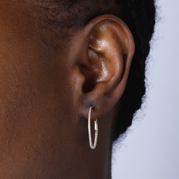 A woman with short black hair is wearing a pair of hoop earrings with a single row of round diamonds.