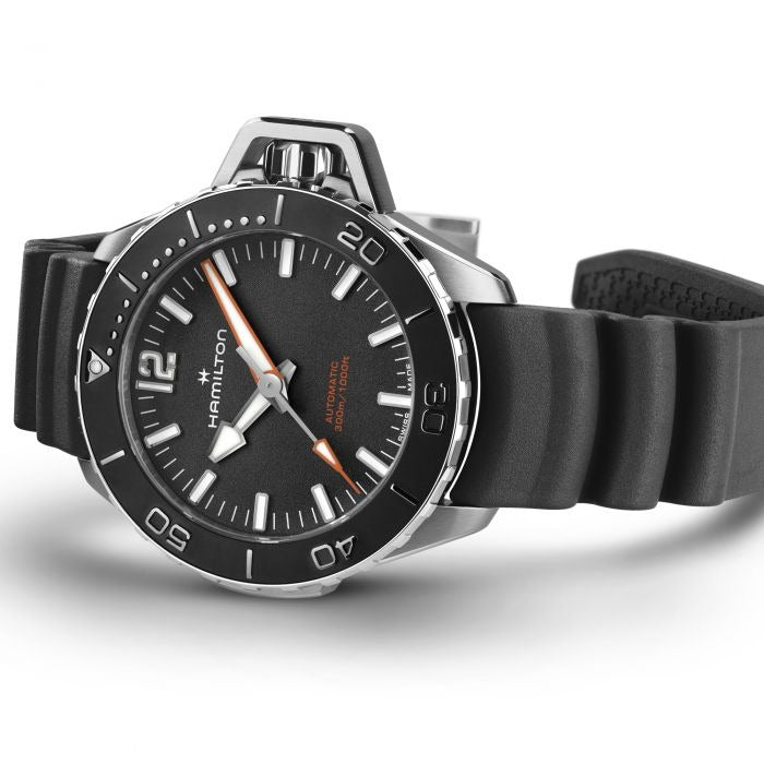 A Hamilton watch is lying on its left side against a white background. It features a black dial, white hands and markers, a steel case, a black bezel, a steel crown, and a black rubber strap.