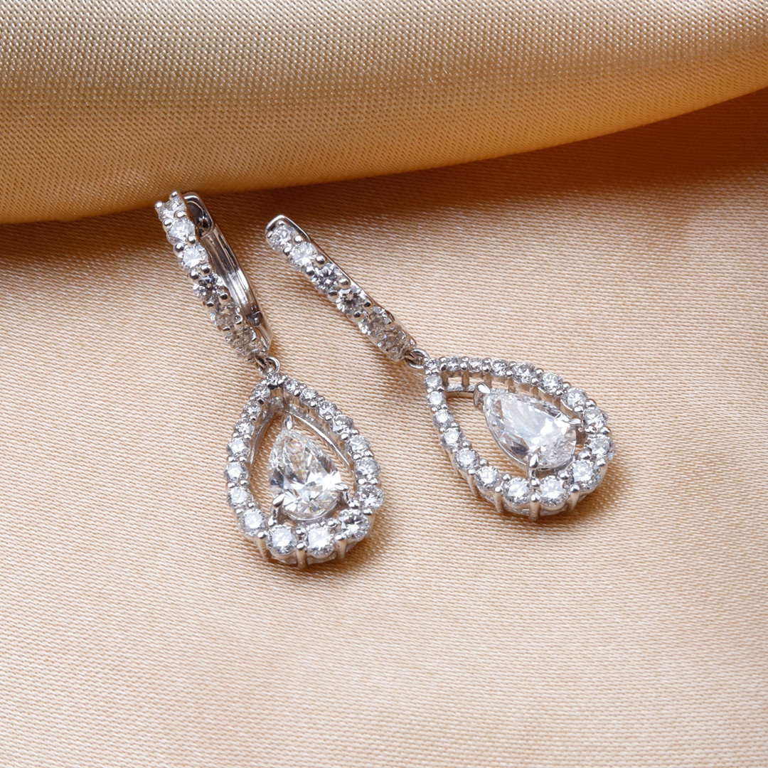 A pair of diamond dangle earrings with a teardrop shaped diamond in the center with a diamond set halo is laying against a gold background.