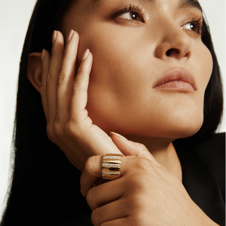 A close-up of a woman wearing a yellow-gold ring. The ring features a wide cigar band with rows of pave-set round diamonds.