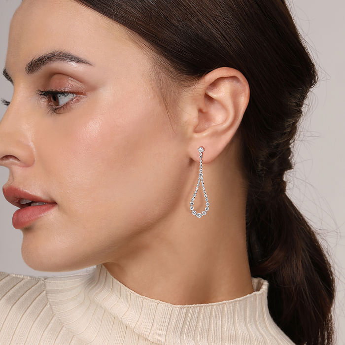 A woman with brunette hair is wearing a white sweater and a pair of drop earrings featuring diamond studs size of the diamond gets bigger towards the bottom.