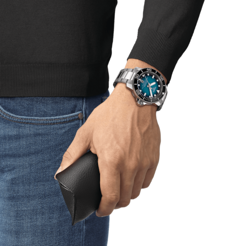 A man wearing a Tissot watch around his wrist. The watch features a black & blue dial, a black bezel, white hands and markers, and a stainless steel bracelet.