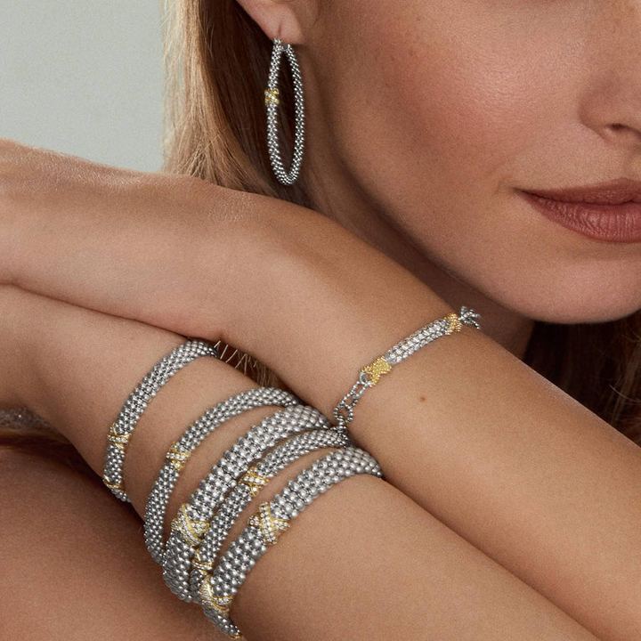 A woman is wearing multiple bracelets and a pair of hoop earrings with a diamond and 18K gold x station with sterling silver caviar beading.