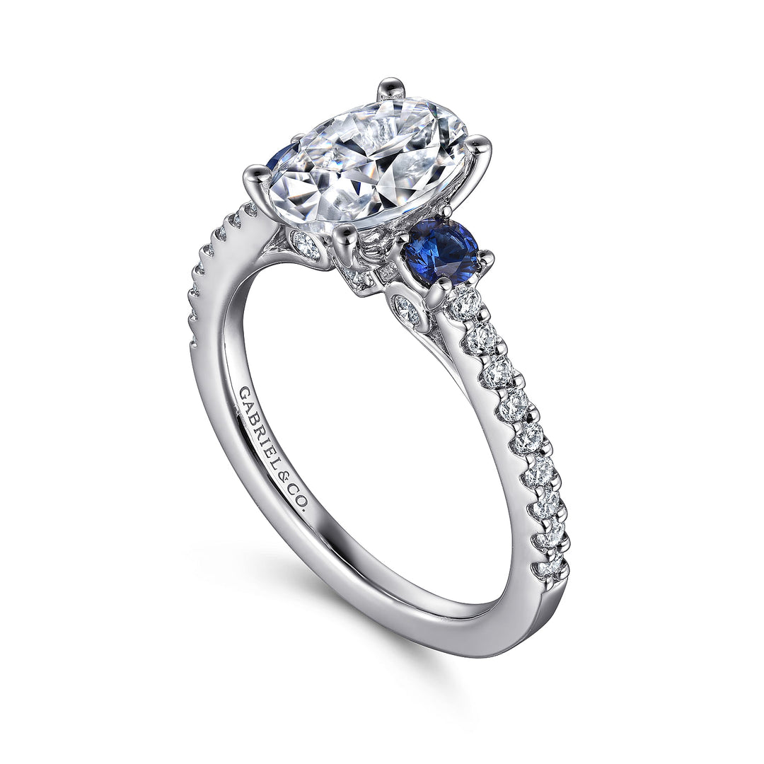 A close-up of a ring made of white gold, angled to the left against a white background. It features a brilliant-cut diamond set in the center, sapphire side stones, and a diamond-set band.