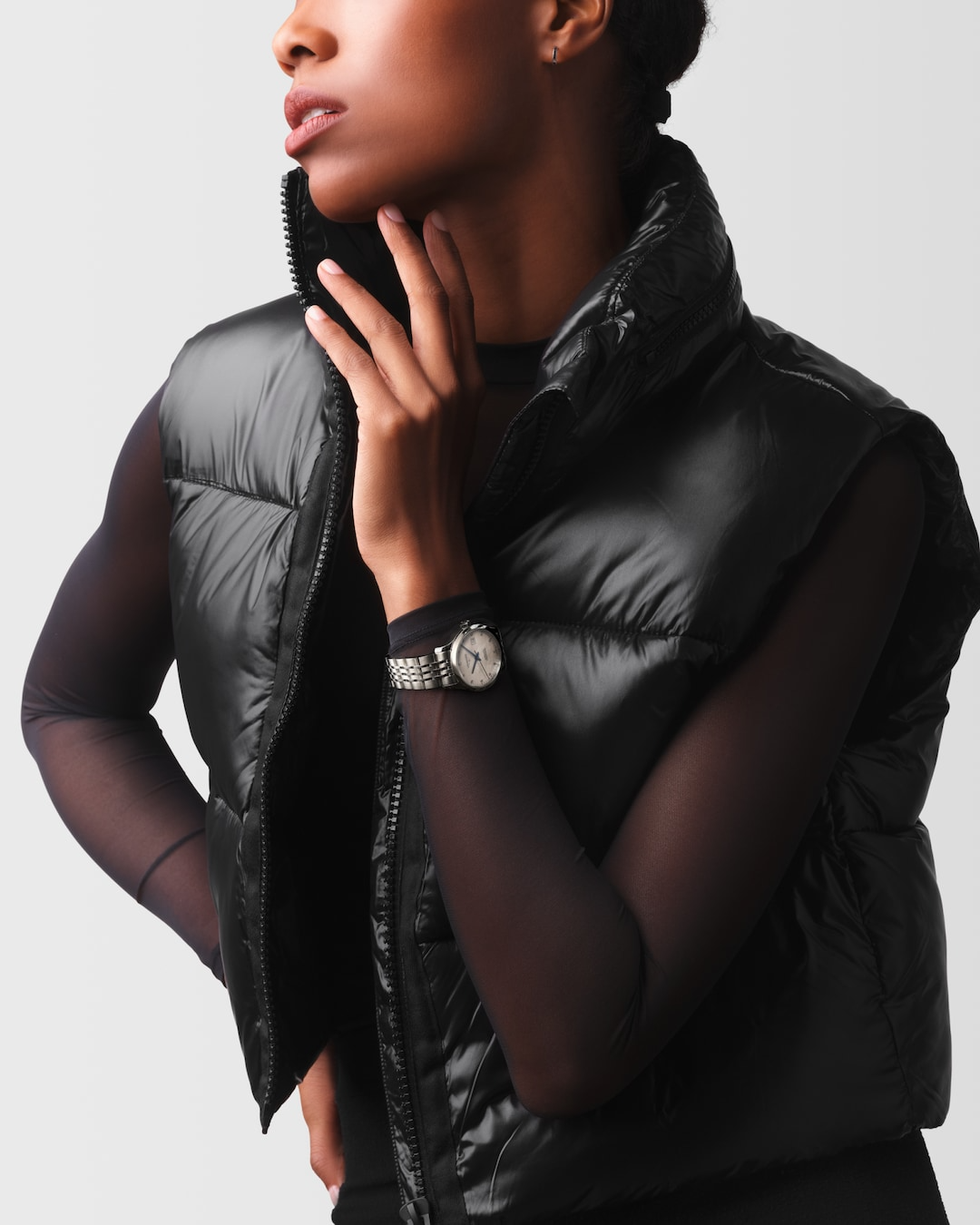 A woman wearing a black puffer vest and a Longines watch around her wrist. The watch features a mother-of-pearl diamond-set dial, blue hands, black markers, a stainless steel bezel, and a stainless steel bracelet.