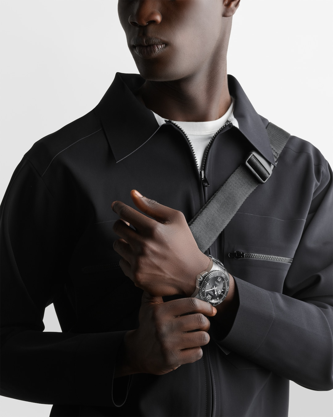 A close-up of a man holding his hand up to his chest with a watch around his wrist. The watch features a black dial, white hands and markers, a black bezel, a crown on the right, and a stainless steel bracelet.