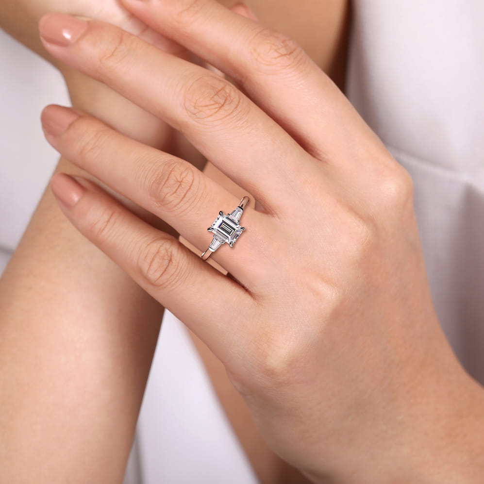 Emerald cut three stone engagement ring worn on a hand, showing the white gold band and baguette side diamonds.