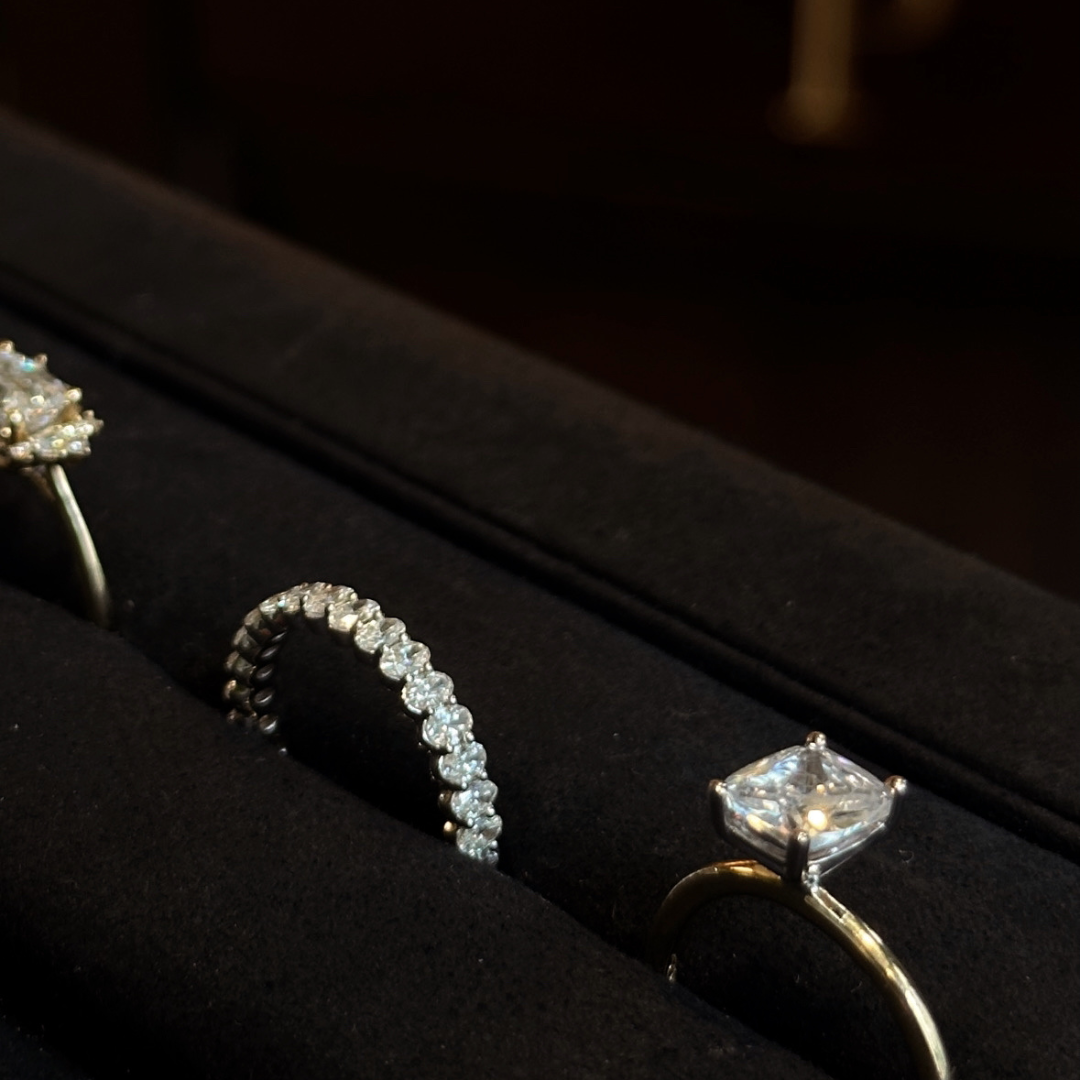 Three rings displayed in a black ring box, including a princess-cut solitaire engagement ring, a diamond-accented band, and a silver-toned floral cluster ring.