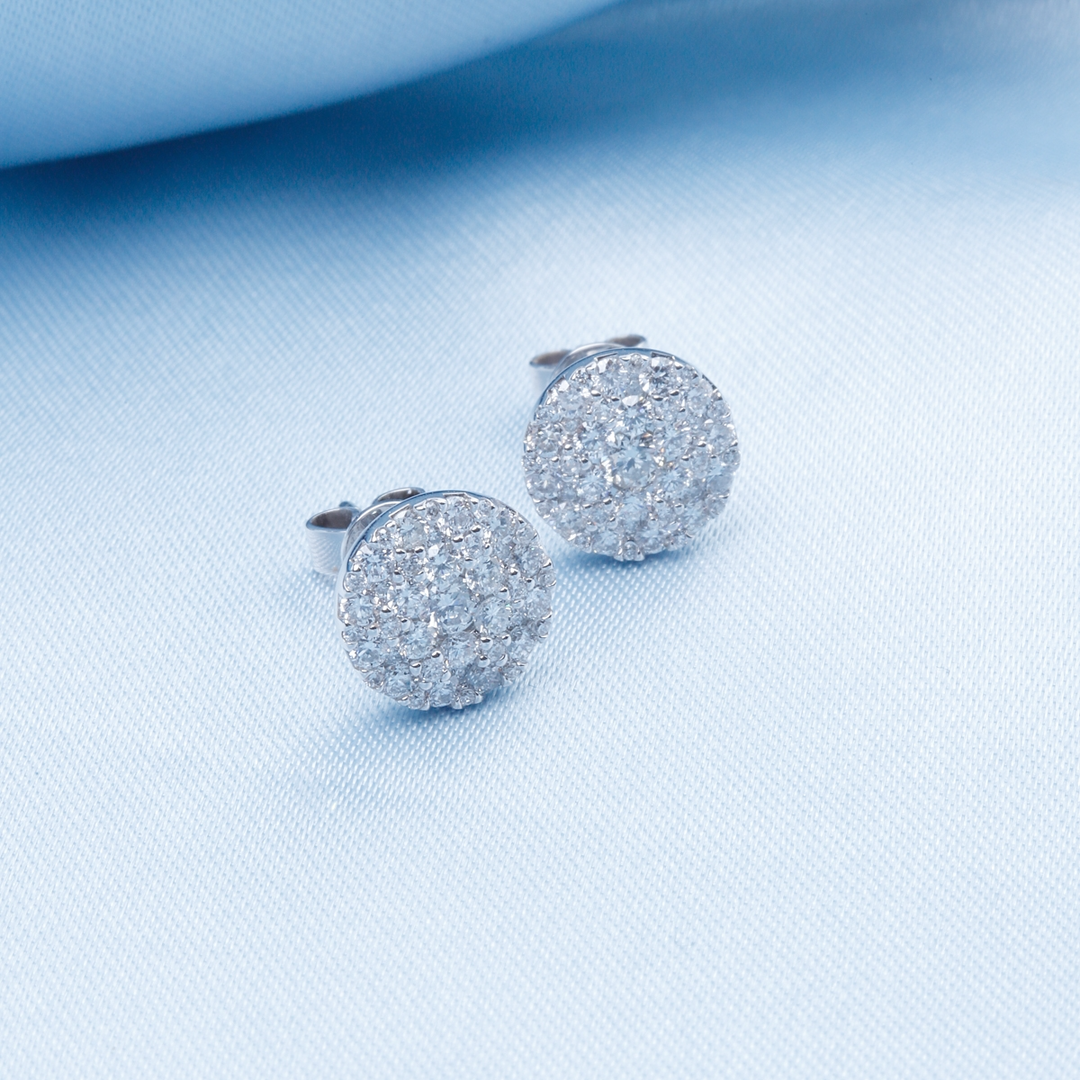 A pair of stud earrings with a round diamond shape with a cluster of diamonds resting on a light blue background.