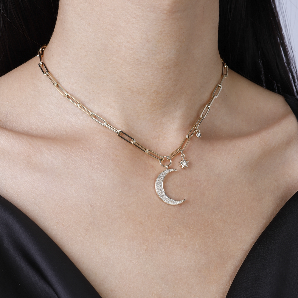 A woman is wearing a yellow-gold Moon and Stars Paperclip Chain and a blue blouse. The necklace features a moon-shaped pendant, 2 round diamonds, and a yellow-gold paperclip chain.