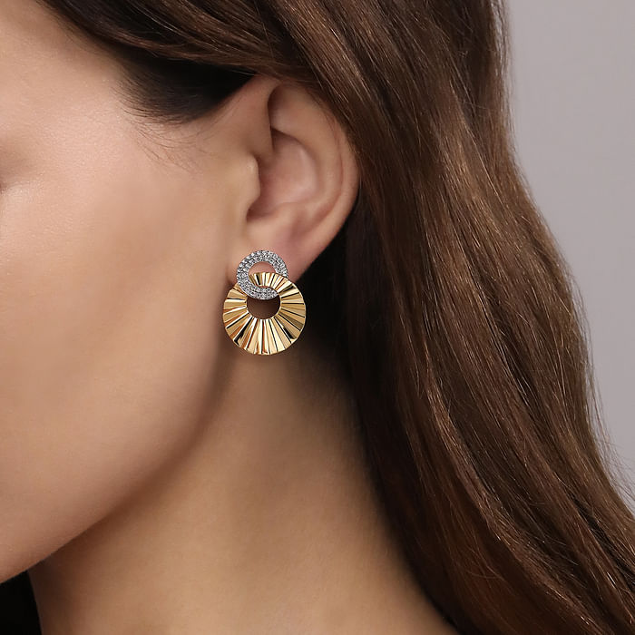 A woman with long brunette hair is wearing a pair of drop earrings with a circle diamond set diamond with an intertwined waved designed gold circle.