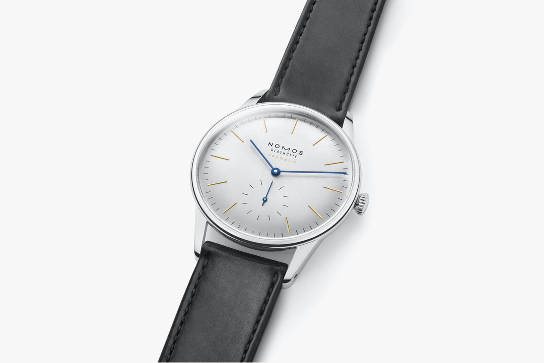 A close-up image of a Nomos Glashutte watch angled to the right. It features a white dial, gold & blue hands and markers, a stainless steel bezel, and a black strap.