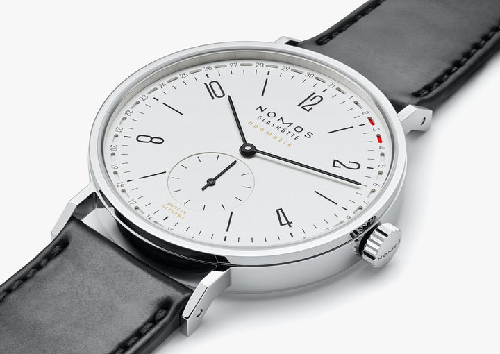 A Nomo Glashutte watch is lying flat and showcasing the dial. It features a white dial, black hands and markers, a stainless steel bezel, and a black leather strap.