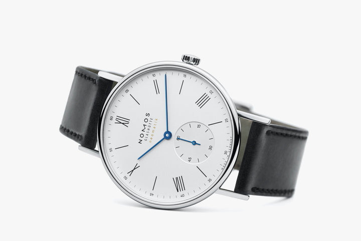 A Nomos Glashutte watch resting on the left side against a white background. It features a white dial, blue hands and markers, a stainless steel bezel, and a black strap.