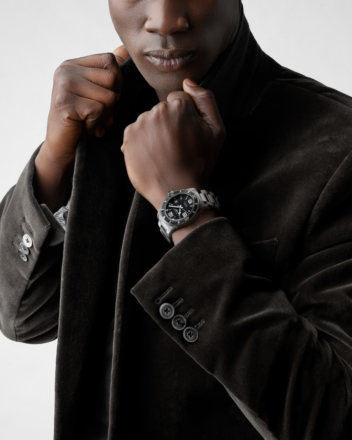 A close-up of a man holding his hand to his jacket with a watch around his wrist. The watch features a black dial, white & silver hands and markers, a black bezel, a crown on the right side, and a stainless steel strap.