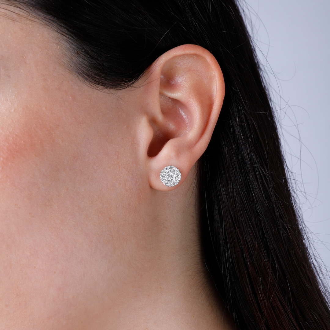 A woman with long brunette hair is wearing a pair of stud earrings with a round diamond shape with a cluster of diamonds. 