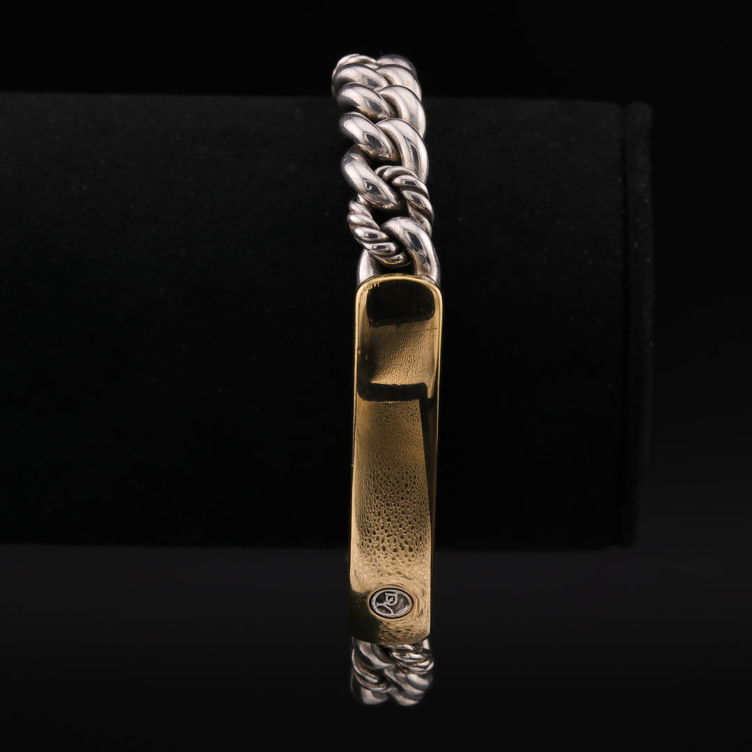 A close-up of a two-toned chain bracelet with a sterling silver curb link bracelet featuring a striking yellow gold ID plate against a black background.