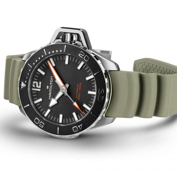 A Hamilton watch is lying on its left side against a white background. It features a black dial, white hands and markers, a steel case, a black bezel, a steel crown, and a green rubber strap.