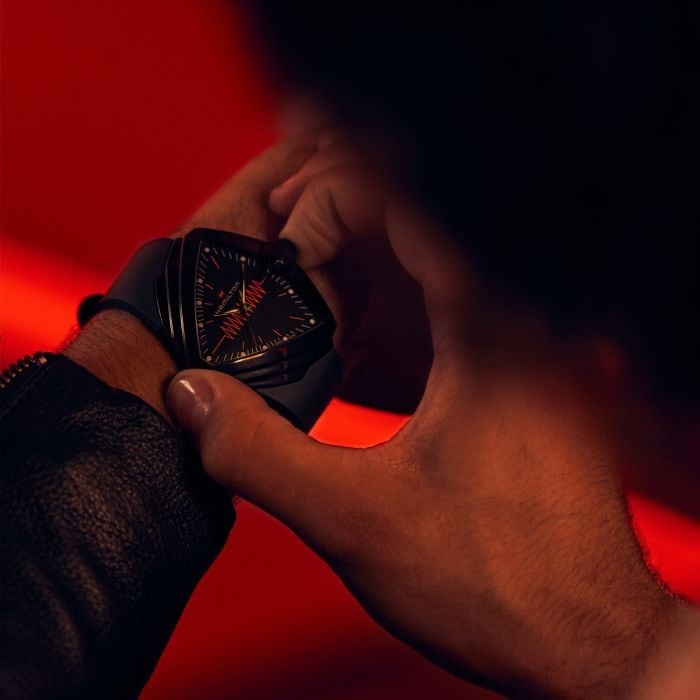 A man is wearing a Hamilton triangular-shaped watch. It features a black dial, white hands and markers, a steel PVD black coating case, a black bezel, a steel crown, and a black rubber strap.